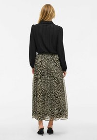 Blouse noire à manches longues avec un motif texturé, associée à une jupe maxi à imprimé léopard, dotée d'un voile transparent et d'une taille froncée. Talons noirs visibles.
