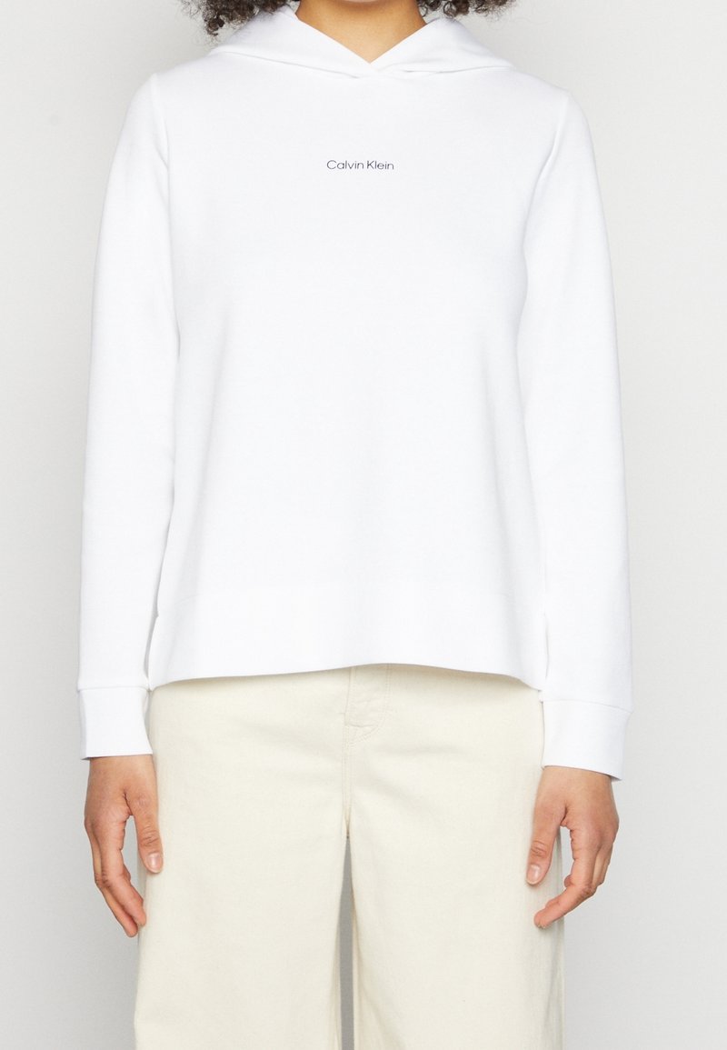 Sudadera blanca con capucha y ajuste relajado, con un pequeño logotipo negro "Calvin Klein" centrado. Tela suave y puños acanalados.