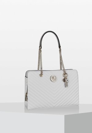 Weiße gesteppte Handtasche mit goldenen Kettenriemen, rundem Logo-Emblem und dekorativen goldenen Anhängern auf einem weißen und grauen Podest.