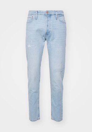 Beige bomber dzseki, kockás ing, világoskék, enyhén megkopott farmernadrág és fekete részletekkel ellátott fehér sneaker. Piros sapka logóval.