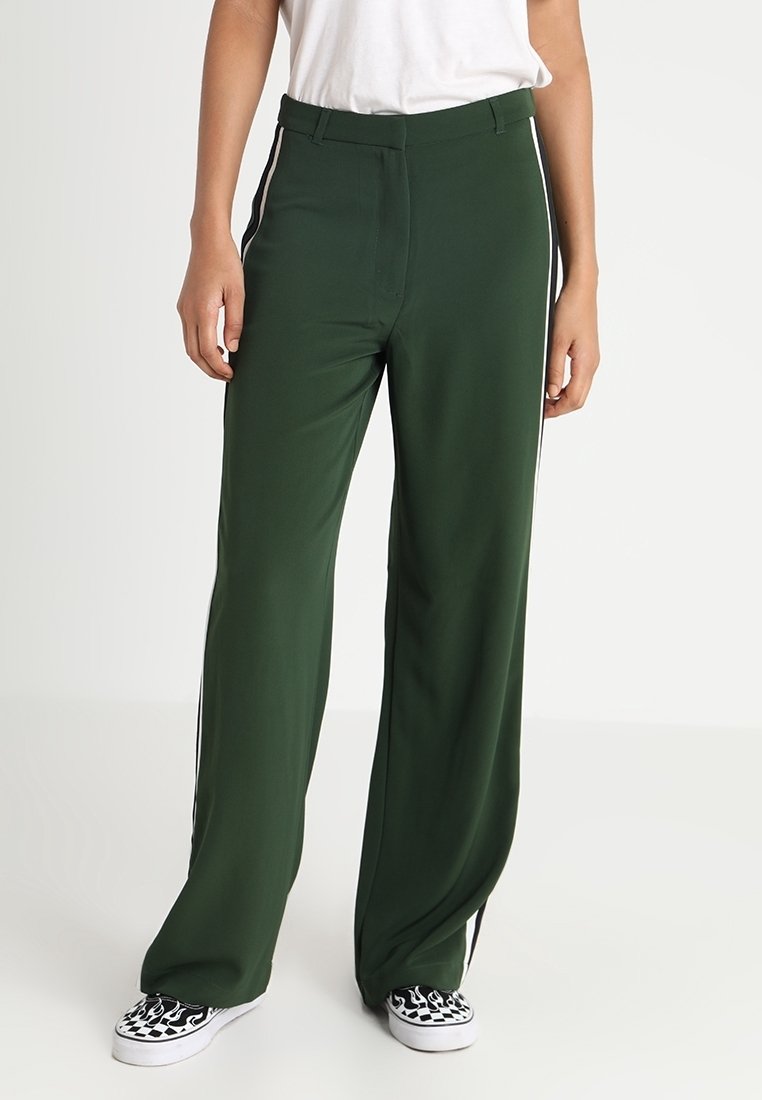Personne portant un pantalon ample vert foncé avec des bandes blanches sur les côtés, des baskets blanches avec des flammes noires, et une chemise blanche.