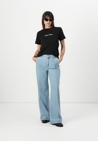 Zwarte katoenen T-shirt met het "Calvin Klein" logo, gecombineerd met lichtblauwe, wijde spijkerbroek. Het model draagt zwarte zonnebril en zwarte schoenen.