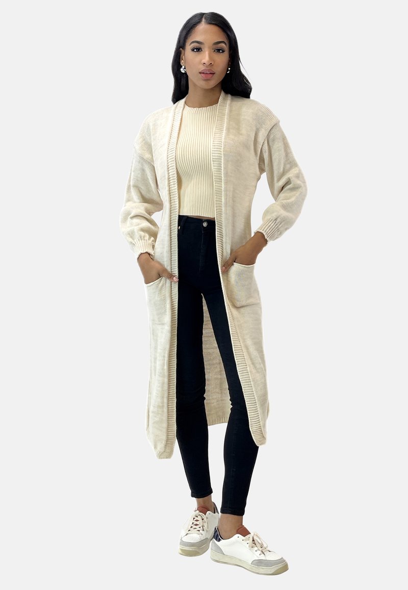 Cardigan lungo beige in morbido tessuto tricot, con bordi a costine, davanti aperto e due tasche anteriori, abbinato a un top aderente crema e pantaloni neri.