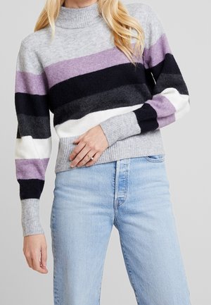 Frau trägt einen gestreiften Pullover mit grauen, violetten, schwarzen und weißen Streifen, kombiniert mit hellblauen Jeans, Hand auf der Hüfte ruhend.