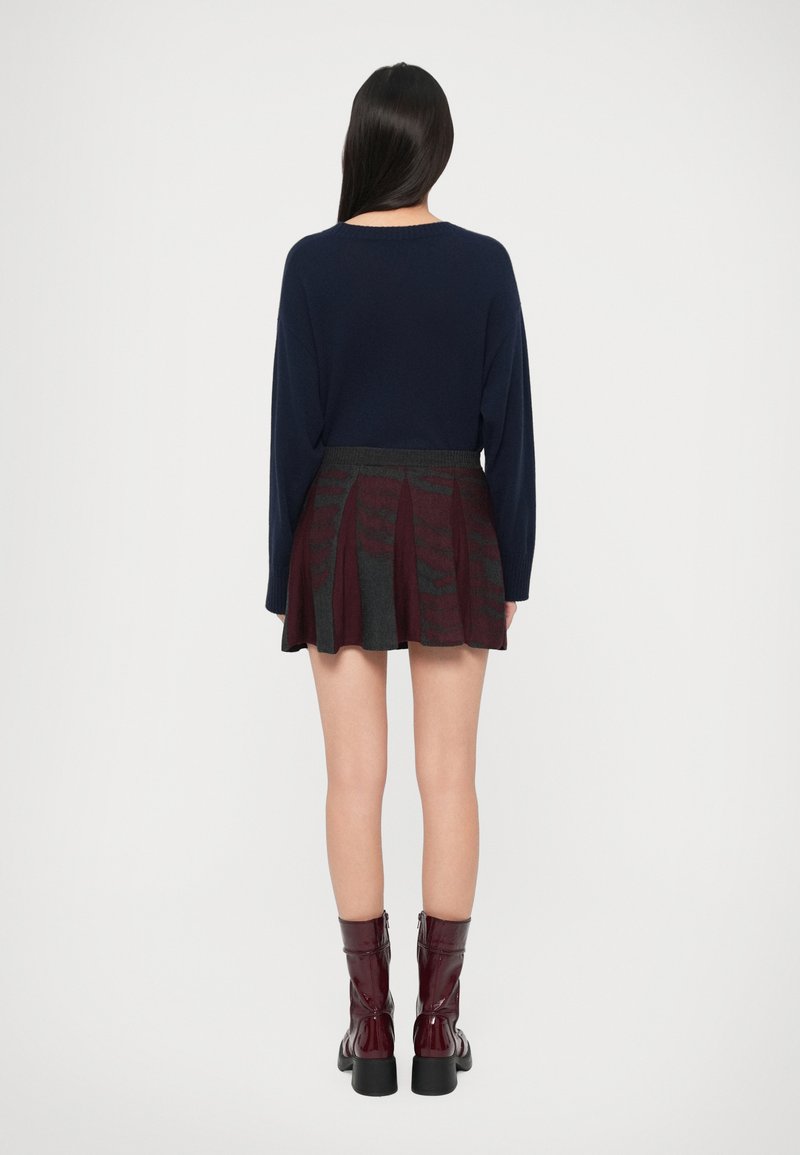 Vrouw met lang donker haar, gekleed in een marineblauwe trui, een geplooide gemusterde rok en donkerrode enkellaarsjes met hoge hakken, staand tegen een effen achtergrond.
