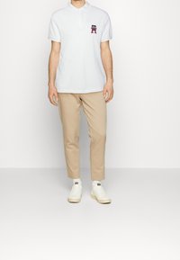 Vit bomullspolo med korta ärmar och en treknäppning; har en färgad logotyp på bröstet. Kombinerad med beige byxor och vita sneakers.