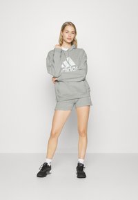 Siva kapuca z dodatnim velikim belim Adidas logotipom, v kombinaciji s sivimi kratkimi hlačami in črnimi športnimi čevlji. Preprost dizajn in udobno prileganje.