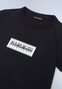 Melna kokvilnas t-krekls ar balto taisnstūra logotipu, kurā ir teksts "NAPAPIJRI geographic". Arapa dizains un īsām piedurknēm.