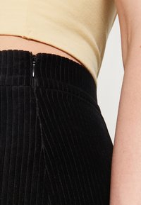 Falda de pana negra con canalones verticales, que presenta una cremallera lateral y una cintura alta, combinada con una blusa ajustada de color beige.