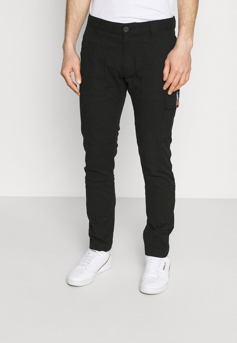 Homme portant un pantalon cargo noir avec poche latérale et des baskets blanches, debout sur un fond uni clair, torse non visible.