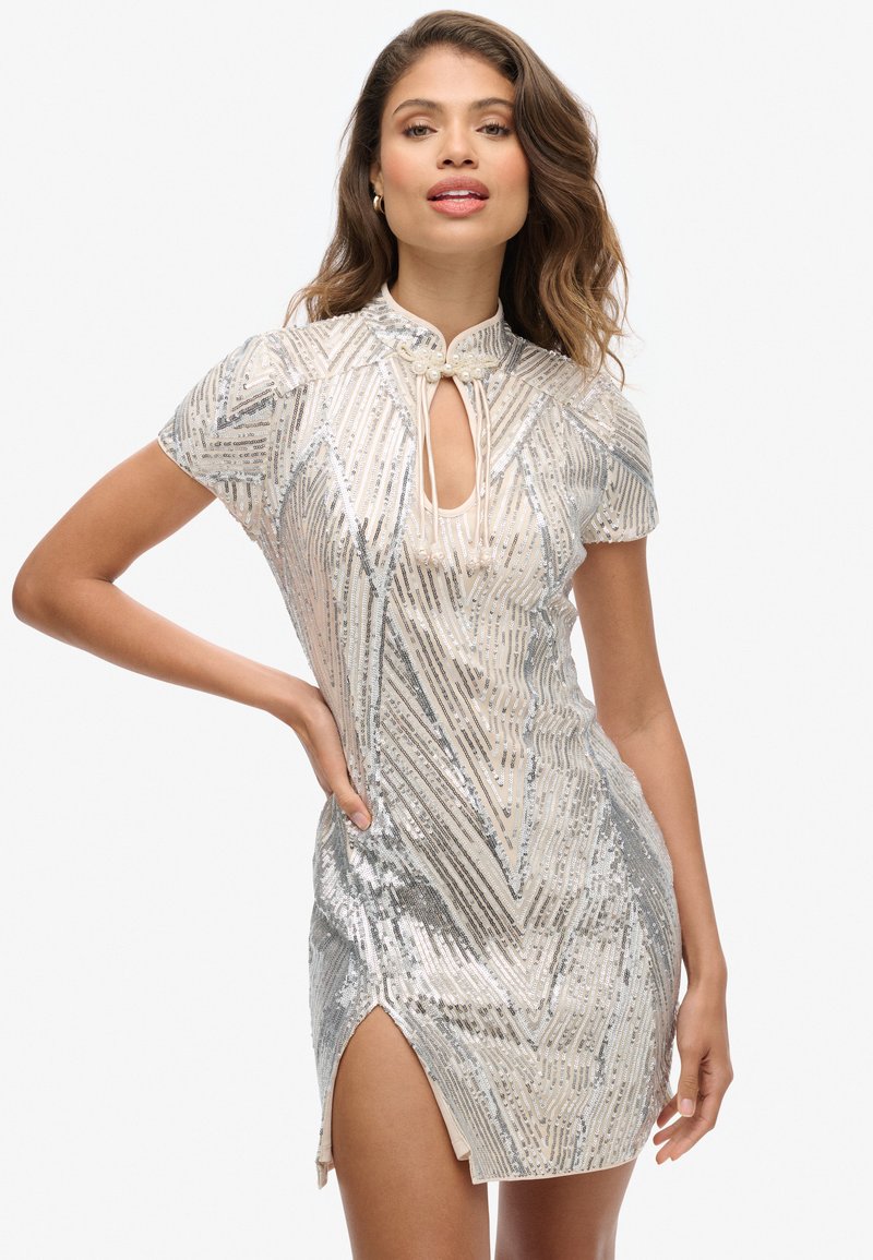 Robe courte en sequins argentés avec un col haut, un décolleté en forme de clé et une fente sur le côté. Présente un motif géométrique et une silhouette ajustée.