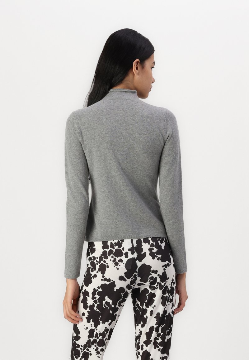 Hellgrauer Pullover mit hohem Kragen und langen Ärmeln, aus weichem Material. Kombiniert mit schwarz-weißen gemusterten Hosen.