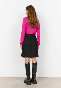 Haut rose à manches longues associé à une jupe noire texturée arrivant au genou et des bottes noires à talons hauts, vu de dos.
