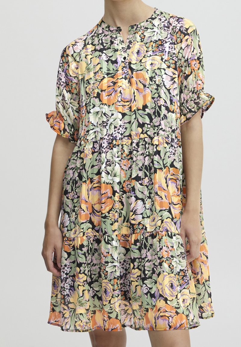 Femme portant une robe fleurie ample à manches courtes avec des fleurs jaunes, oranges et violettes sur un fond sombre.