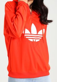 Red pullover hoodie with a half-zip, featuring a large white Adidas logo. Made from smooth material, it has a front kangaroo pocket and ribbed cuffs.