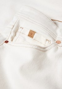 Tejido de denim blanco con un bolsillo que exhibe una pequeña etiqueta de cuero marrón. Acentos de hardware en color cobre y detalles de costura en contraste visibles.