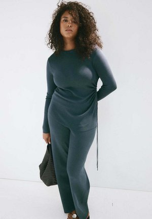 Mujer con cabello rizado lleva un top de manga larga y pantalones gris oscuro, sosteniendo un bolso negro texturizado contra un fondo claro y sencillo.