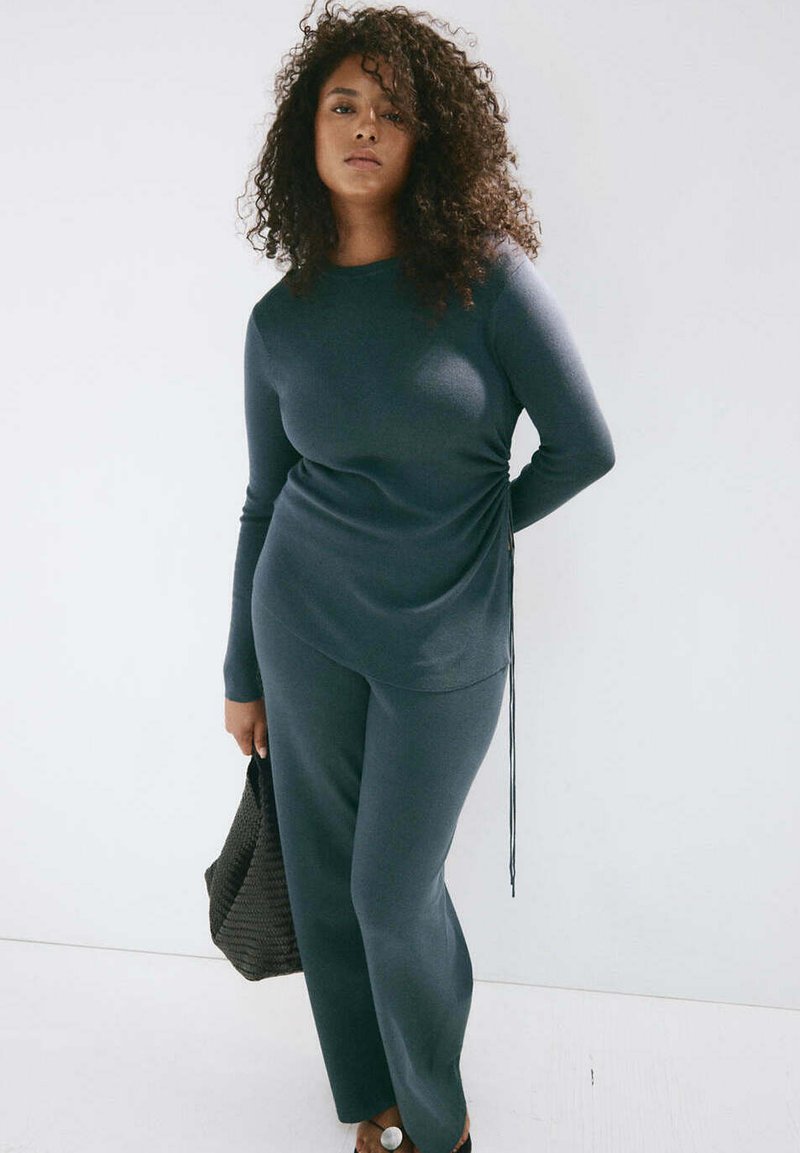Femme aux cheveux bouclés portant un haut à manches longues gris foncé et un pantalon, tenant un sac à main noir texturé sur un fond clair uni.