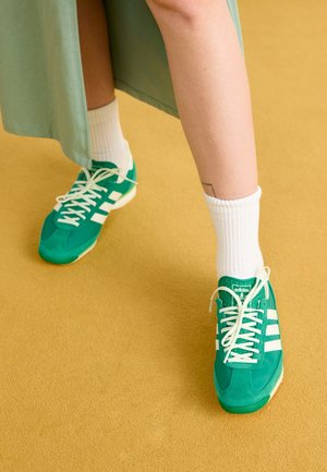 Osoba nosząca zielone adidasy Adidas z białymi paskami, białe skarpety i zieloną spódnicę stojąca na podłodze koloru musztardowego.