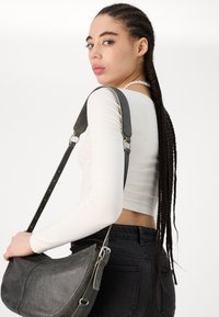 Bolsa de ombro em pele preta com um formato curvo, ferragens prateadas e alça ajustável, usada com uma blusa justa de manga longa branca e jeans pretos.