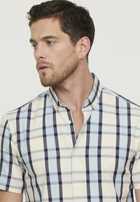 Jeune homme aux cheveux bruns courts portant une chemise à manches courtes à carreaux bleu, crème et marine, regardant vers sa gauche.