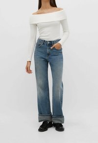 Biała, prążkowana bluzka na jedno ramię w połączeniu z wysoko uszytymi, szerokimi niebieskimi dżinsami, z podwiniętymi mankietami i klasyczną fakturą dżinsu. Czarne buty.