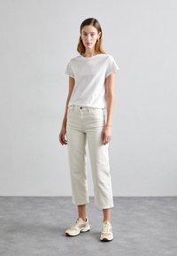 Mladá žena se rovnými hnědými vlasy má na sobě bílé tričko, krémové zkrácené džíny a béžovo-bílé tenisky, stojí před jednobarevným šedým pozadím.