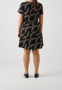 Zwarte jurk met crèmekleurig bladrandpatroon, korte mouwen, getailleerde taille, uitlopende rok, gemaakt van soepele stof. Gedragen met zwarte schoenen.