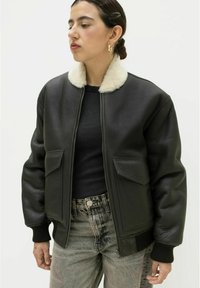 Schwarze Lederbomberjacke mit einem cremefarbenen Kunstfellkragen, gerippten Bündchen und zwei Fronttaschen. Getragen über einem schwarzen Rippoberteil und gemusterten Jeans.
