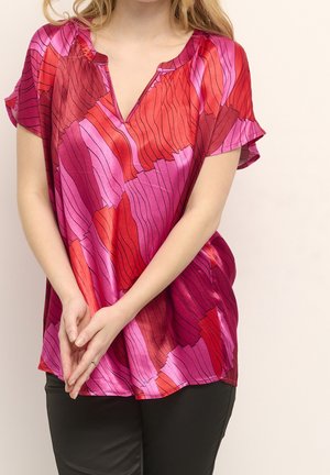 Frau trägt eine kurzärmelige, lockere Bluse mit roten und pinken abstrakten Wellenmustern, kombiniert mit schwarzen Hosen, steht mit verschränkten Händen.