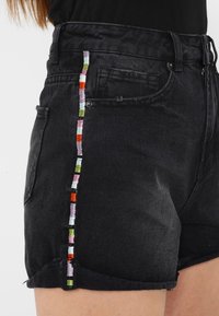 Mustat farkkushortsit, joissa on frangeerattu helma ja värikkäitä brodeerattuja yksityiskohtia sivuilla, mukaan lukien vaaleanpunaisia, vihreitä ja oransseja lankoja.