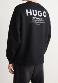 Persona che indossa una felpa nera con la scritta bianca "HUGO MCMXCIII Marchio Registrato Internazionalmente Fondato nel 1993" sulla schiena.