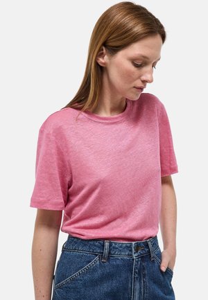 Femme aux cheveux bruns raides portant un t-shirt rose à manches courtes rentré dans un jean bleu taille haute, regardant vers le bas sur sa gauche.