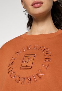 Terrakotta színű pulóver kör alakú, kiemelt hímzett "Nike Court" logóval. Kerek nyakú dizájn, puha textúrával és minimalista stílussal.