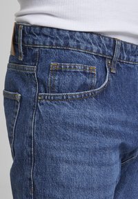 Prim-plan cu blugi din denim albastru purtați cu o cămașă albă reiată băgată în pantaloni, arătând buzunarul frontal și buclele pentru curea.