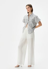 Camicia a righe con maniche corte, due tasche frontali, abbinata a pantaloni ampi e trasparenti bianchi e sandali con apertura davanti.