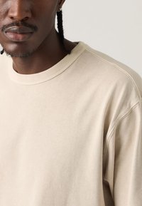 Κοντινό πλάνο ενός ατόμου με πλεξούδες που φοράει μπεζ φούτερ με στρογγυλή λαιμόκοψη πάνω σε απλό φωτεινό φόντο.