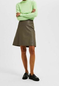 Top a maniche lunghe verde a coste abbinato a una gonna A-line in pelle marrone e mocassini neri. Focus sulla rigatura, sulla texture liscia della pelle e sul taglio.