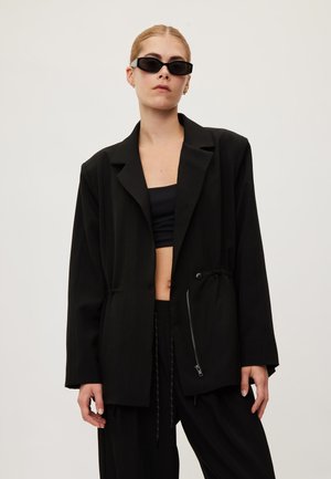 Femme portant un blazer noir oversize, un crop top noir, un pantalon assorti et des lunettes de soleil noires, debout devant un fond clair uni.