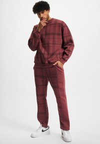 Maronfarbener Sweatshirt- und Hosenanzug mit schwarzem Karomuster, gerippten Bündchen und Seitentaschen. Getragen mit weißen Sneakern.