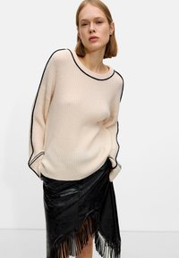 Suéter beige de canalé con ribete negro, acompañado de una falda de cuero negra con un dobladillo de flecos. El conjunto combina texturas suaves con un diseño estructurado.