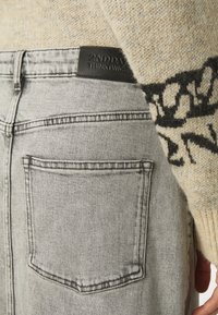 Jeans en denim gris avec une poche arrière, arborant une étiquette en cuir noir sur la taille avec du texte en relief. Pull en beige clair avec un motif foncé.