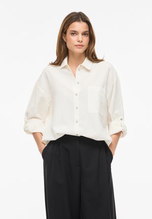 Žena s hnědými vlasy, oblečená v volné bílé košili s vyhrnutými rukávy a černých širokých kalhotách, stojí s rukama v kapsách.