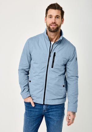 Man wearing light blue zip-up jacket and blue jeans, standing with one hand in pocket against plain background.