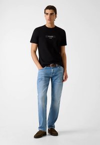 Schwarzes Baumwoll-T-Shirt mit "GUESS"-Logo, hellblaue Jeans aus Denim und braune Mokassins aus Wildleder. Model steht vor neutralem Hintergrund.