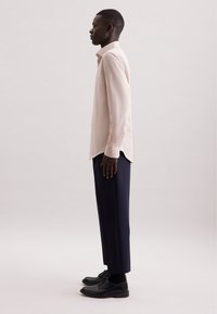 Camicia a maniche lunghe rosa chiaro in tessuto texturizzato, abbinata a pantaloni blu navy e scarpe nere, vista di lato su uno sfondo neutro.