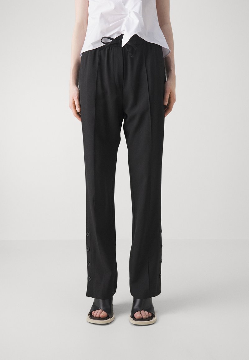 Black trousers with a relaxed fit, featuring a drawstring waist and side buttons along the hem, paired with black slide sandals.