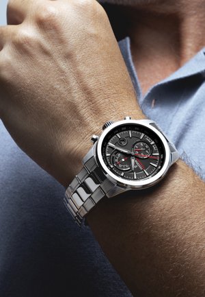 Montre-bracelet en métal argenté avec un cadran noir, comportant trois sous-cadrans et des accents rouges. Le bracelet a une finition polie avec des maillons rectangulaires.