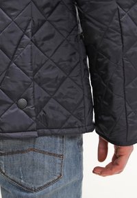 Nahaufnahme einer Person, die eine schwarze Steppjacke und blaue Jeans trägt. Es wird der untere Rücken der Jacke und die rechte Hand gezeigt.