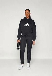 Sweatshirt preto da Adidas com logotipo branco, combinado com calças de treino pretas ajustadas. Modelo segura uma botija de água, usando ténis brancos. Tecido suave e casual.